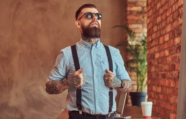 Bir gömlek ve pantolon askısı, içinde güneş gözlüğü takıyor, bir dizüstü bilgisayar ile bir resepsiyon yakın duran, çatı katı iç bir ofiste pencere bakarak eski moda dövmeli hipster.