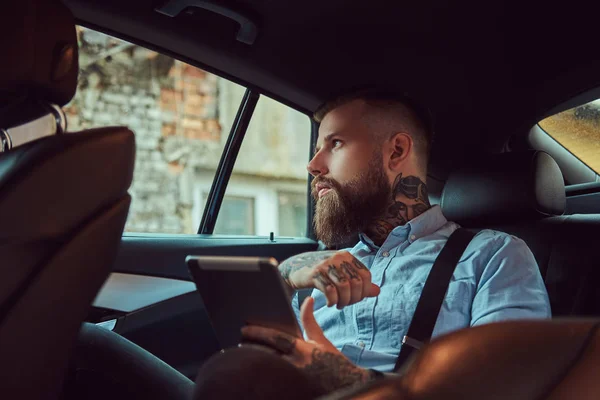 Old-fashioned tattooed hipster guy in a shirt with suspenders, holding a tablet while sitting in a luxury car on a back seat, looking away. — Stock Photo, Image