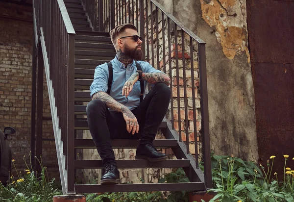 Stylish handsome old-fashioned tattooed hipster guy in a shirt with suspenders, sitting on stairs near old building outdoors. — Stock Photo, Image