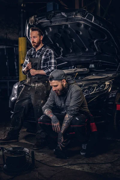Two mechanic sit on a broken car, rest after a hard days work in a garage. — Stock Photo, Image