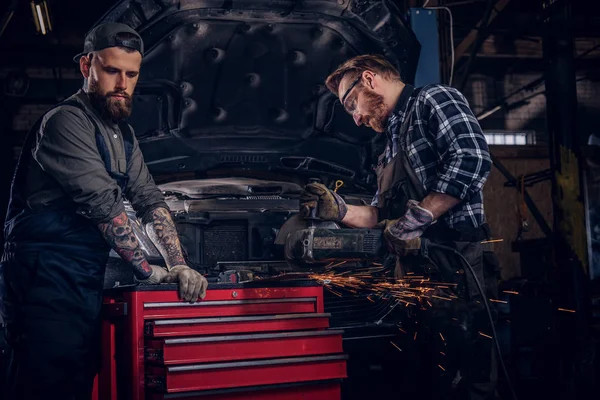 Deux mécaniciens barbus dans un uniforme et des lunettes de sécurité travaillant avec une meuleuse d'angle tout en se tenant contre une voiture cassée dans le garage de réparation . — Photo