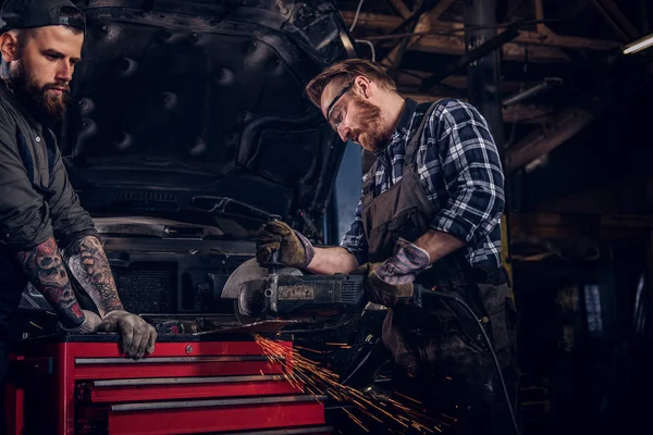 Dwa Brodaty auto mechanik w mundurze i okulary ochronne, praca z szlifierki kątowej stojąc przed uszkodzony samochód w garażu napraw. — Zdjęcie stockowe