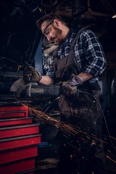 Brutální auto mechanik v uniformě a ochranné brýle práci s úhlovou brusku stoje proti rozbité auto v garáži opravy. — Stock fotografie