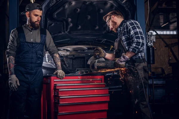 Deux mécaniciens barbus dans un uniforme et des lunettes de sécurité travaillant avec une meuleuse d'angle tout en se tenant contre une voiture cassée dans le garage de réparation . — Photo