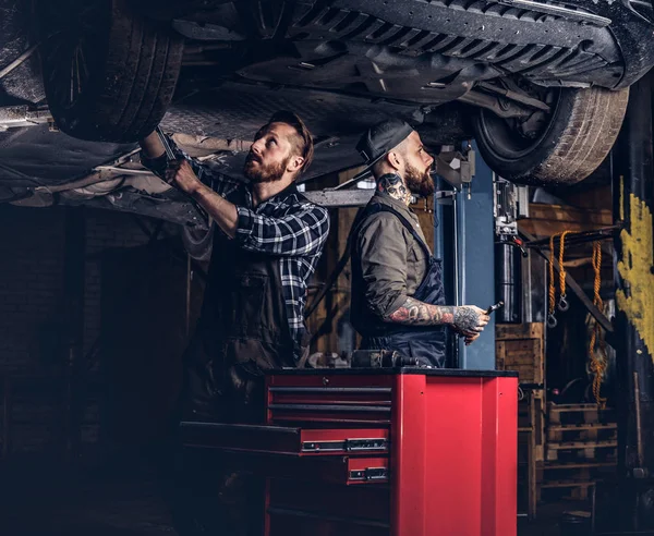 Deux mécaniciens barbus dans un uniforme réparer la suspension des voitures tout en se tenant sous la voiture de levage dans le garage de réparation . — Photo