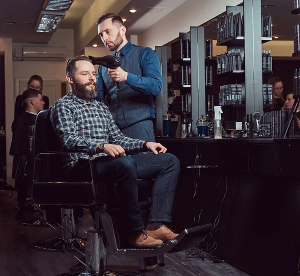 Barbier professionnel travaillant avec un client dans un salon de coiffure, utilise un sèche-cheveux . — Photo