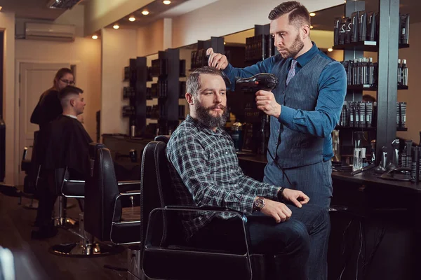 Barbeiro profissional trabalhando com um cliente em um salão de cabeleireiro, usa um secador de cabelo . — Fotografia de Stock