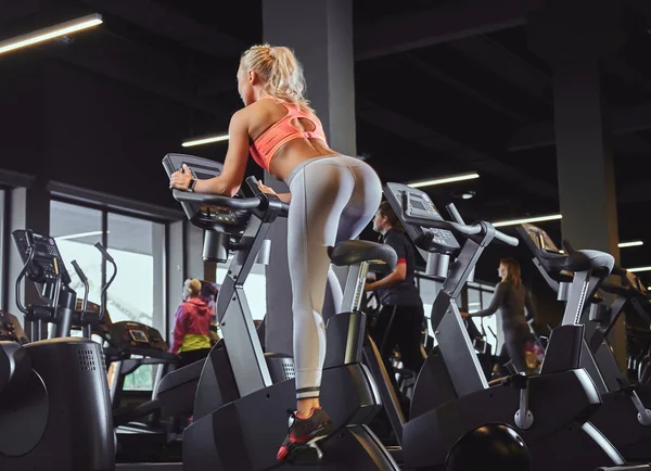 Молода красива блондинка в спортивному одязі тренується на тренажерному велосипеді в сучасному фітнес-центрі . — стокове фото