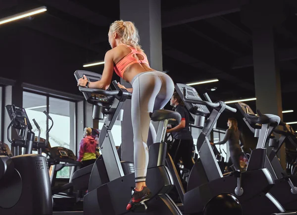 Joven hermosa mujer rubia en entrenamiento de ropa deportiva en una bicicleta estática en el moderno gimnasio . — Foto de Stock