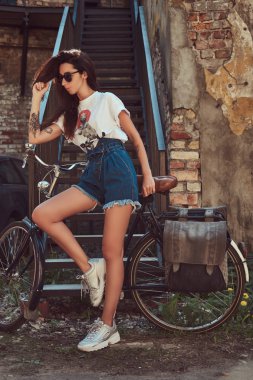 İnce kız kot şort, beyaz tişört ve şehir Bisiklet ile poz güneş gözlüğü, kısacası yakınındaki bir eski terkedilmiş tuğla bina.