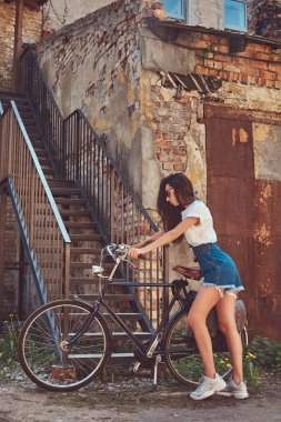 İnce kız kot şort, beyaz tişört ve şehir Bisiklet ile poz güneş gözlüğü, kısacası yakınındaki bir eski terkedilmiş tuğla bina.
