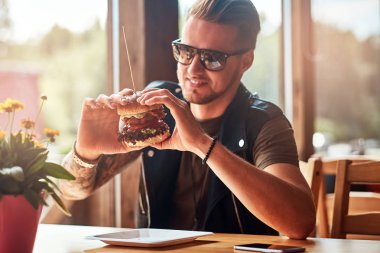 Bir hamburger yeme bir şık saç ve sakal ile yakışıklı hipster yol kenarına yerleştirilen bir kafede yemek yemeye karar verdim bir masada oturur.