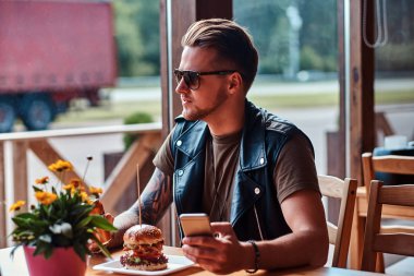 Bir şık saç ve sakal ile yakışıklı hipster yol kenarına yerleştirilen bir kafede öğle yemeği sırasında bir telefon tutarak bir masada oturur.