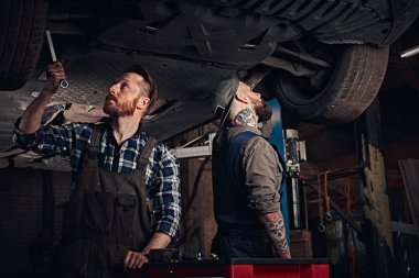 Üniformalı iki sakallı otomatik mekanik tamir onarım garajda araba kaldırma altında duran araçların süspansiyon. 