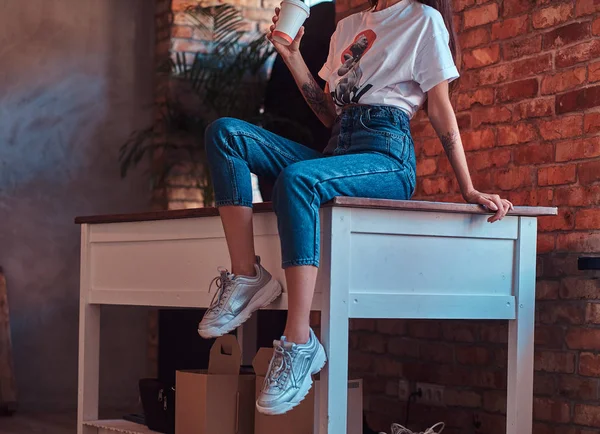 一个凉爽的现代女孩的形象, 穿着白色的顶部和牛仔裤坐在桌子上, 一杯外卖咖啡与阁楼内部的工作室. — 图库照片