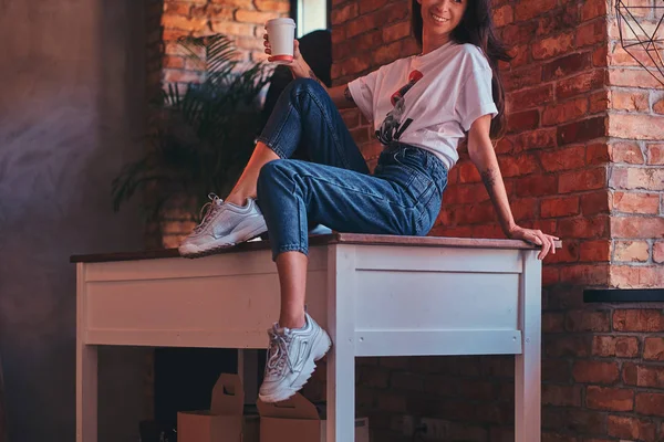 Imagen recortada de una chica moderna y fresca con un top blanco y vaqueros sentados en una mesa con una taza de café para llevar en el estudio con interior loft . — Foto de Stock