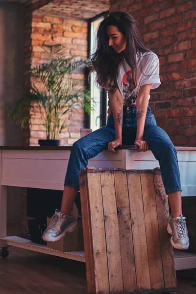 Menina moderna legal vestindo um top branco e jeans sentados em uma mesa em estúdio com interior loft . — Fotografia de Stock