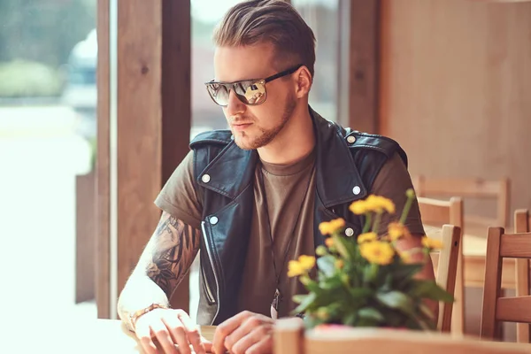 Hipster con un corte de pelo elegante y barba se sienta en una mesa en un café al lado de la carretera . — Foto de Stock