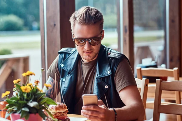 Hipster με μοντέρνο κούρεμα και γενειάδα που κάθεται σε ένα τραπέζι, χρησιμοποιώντας ένα smartphone κατά τη διάρκεια γεύματος σε οδική καφέ. — Φωτογραφία Αρχείου