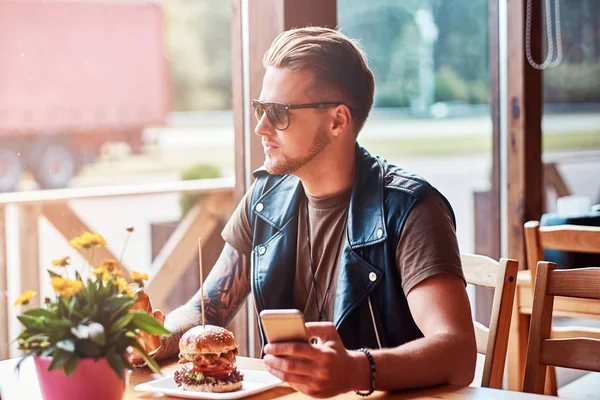 Красивый хипстер со стильной стрижкой и бородой сидит за столом, держа телефон во время обеда в придорожном кафе . — стоковое фото