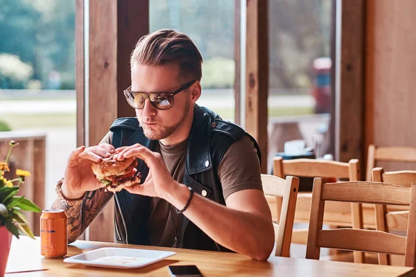 Красавчик-хипстер со стильной стрижкой и бородой сидит за столом, решил пообедать в придорожном кафе, съесть гамбургер . — стоковое фото