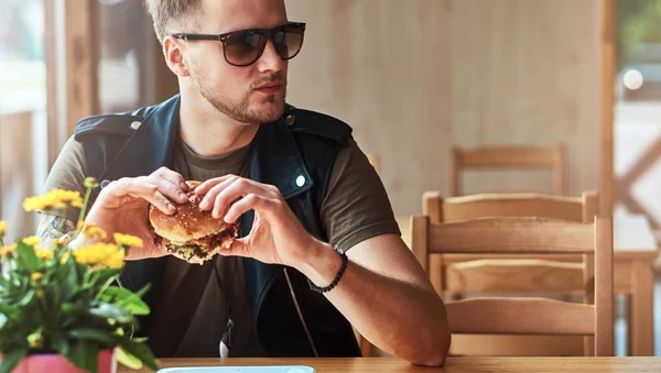Красавчик-хипстер со стильной стрижкой и бородой сидит за столом, решил пообедать в придорожном кафе, съесть гамбургер . — стоковое фото