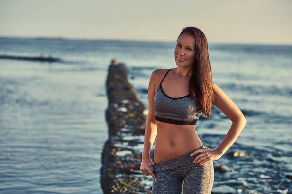 Mooie fitness vrouw in een grijze sportkleding poseren tegen een achtergrond van de zee. — Stockfoto