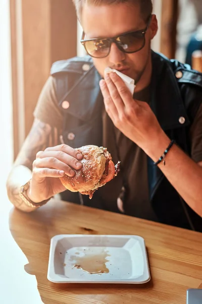 Beau hipster avec une coiffure élégante et barbe assis à une table, a décidé de dîner dans un café en bord de route, en mangeant un hamburger . — Photo