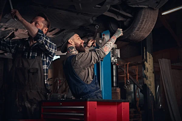 Deux mécaniciens barbus dans un uniforme réparer la suspension des voitures tout en se tenant sous la voiture de levage dans le garage de réparation . — Photo