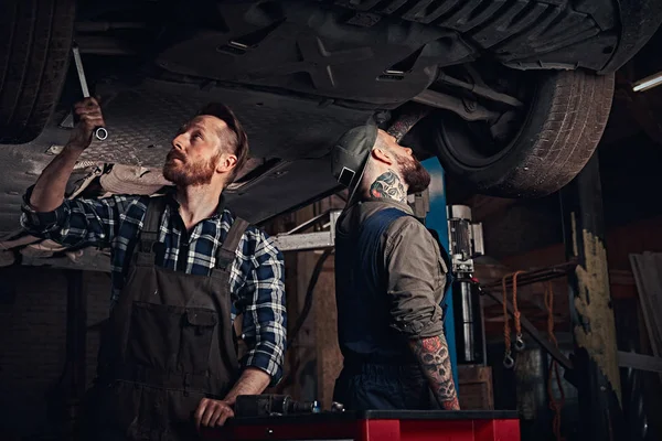 Dwóch brodatych auto mechanik w mundurze naprawy zawieszenia samochodów, stojąc pod podnoszenia samochodu w garażu napraw. — Zdjęcie stockowe