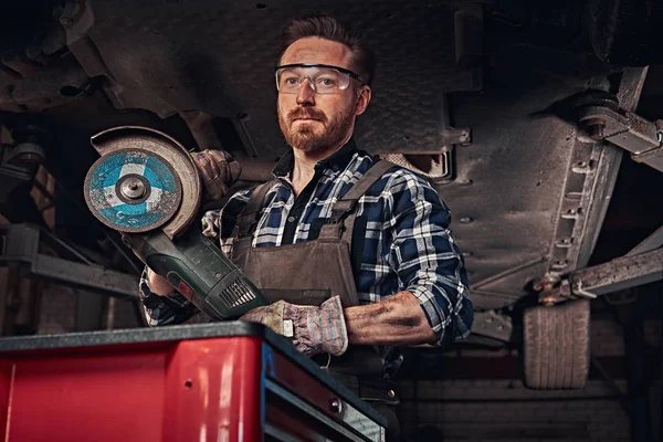 Mechanic in a uniform and safety glasses holds an angle grinder while standing under lifting car in a repair garage. — Stock Photo, Image