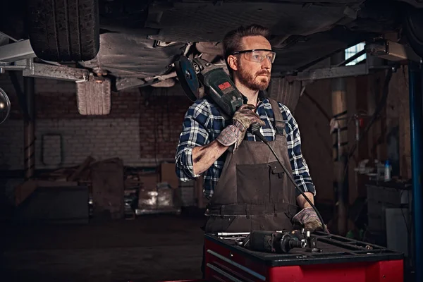 Mécanicien dans un uniforme et lunettes de sécurité tient une meuleuse d'angle tout en se tenant sous la voiture de levage dans un garage de réparation . — Photo