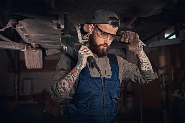 Mecánico en un uniforme y gafas de seguridad sostiene una amoladora angular mientras está de pie debajo de levantar el coche en un garaje de reparación . —  Fotos de Stock
