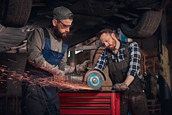 Two bearded auto mechanic in a uniform and safety glasses working with an angle grinder while standing under lifting car in repair garage.