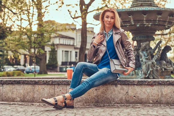 Модная блондинка расслабляет на открытом воздухе, пьет кофе на вынос, сидя на скамейке в городском парке . — стоковое фото