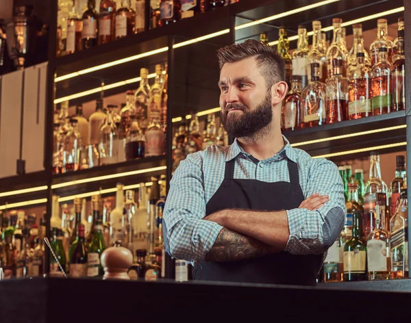 Elegante barista brutale in camicia e grembiule in piedi con braccia incrociate sullo sfondo del bancone del bar . — Foto Stock