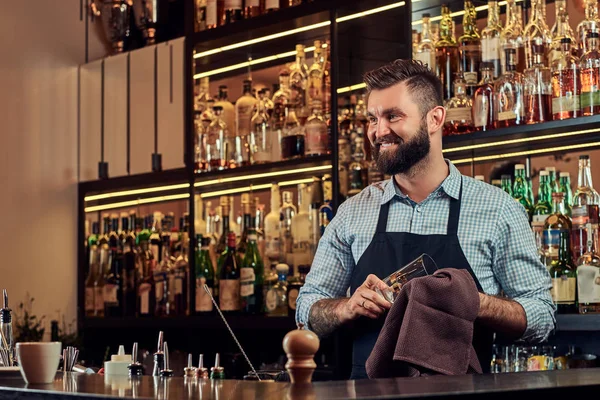 Bartender brutal yang ceria sedang membersihkan kaca dengan kain di latar belakang bar counter . — Stok Foto