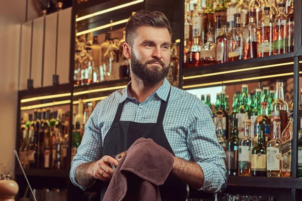 Elegante barman brutal está limpiando el vidrio con un paño en el fondo del mostrador de bar . —  Fotos de Stock