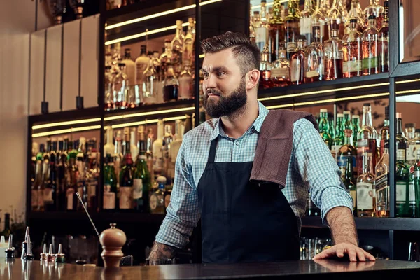Stijlvolle bebaarde barman in een shirt en schort staande op bar teller achtergrond. — Stockfoto