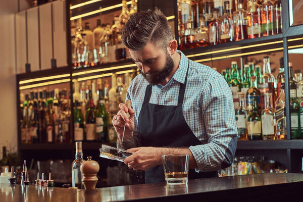 Stylish bearded barman in a shirt and apron splits ice to make a cocktail at bar counter background.