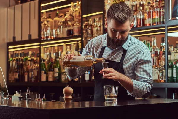 Стильный жестокий бармен в рубашке и фартуке делает коктейль на заднем плане барной стойки . — стоковое фото