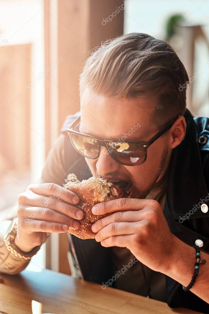 Portrait of a hungry hipster guy with a stylish haircut and beard sits at a table, decided to dine at a roadside cafe, eating a hamburger.