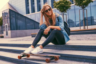 Genç hipster sarışın kız rahat kıyafetler ve adımları bir gökdelen karşı oturup güneş gözlüğü, kaykay üzerinde sürme sonra dinlenme.