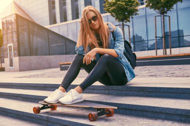 Genç hipster sarışın kız rahat kıyafetler ve adımları bir gökdelen karşı oturup güneş gözlüğü, kaykay üzerinde sürme sonra dinlenme.