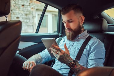 Eski moda dövmeli hipster erkekte bir lüks araba arka koltukta otururken bir tablet kullanarak bir gömlek askısı,.