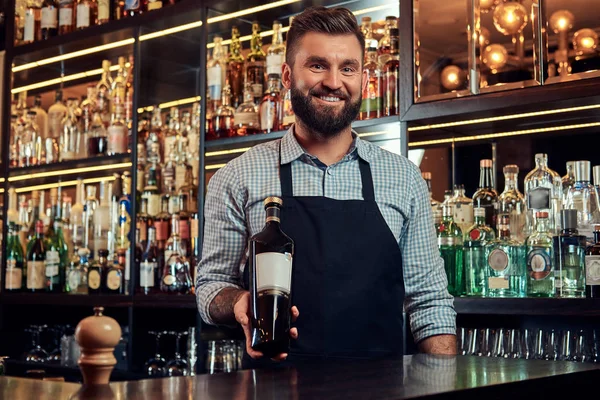 Neşeli şık acımasız barmen bir gömlek ve önlük özel alkol counter arka plan bir şişe sunar. — Stok fotoğraf