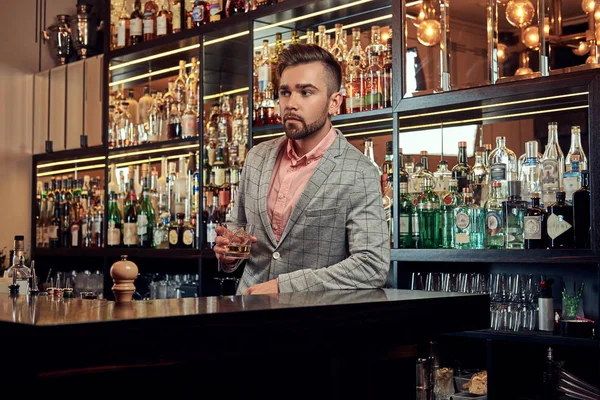 Stylový pohledný muž v elegantním obleku drží sklenku whisky v baru counter pozadí. — Stock fotografie
