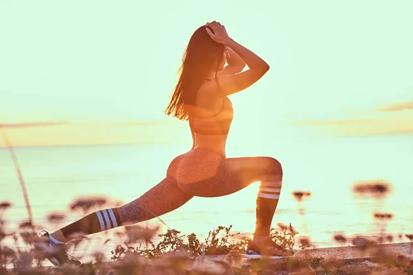 Fitness woman practicing stretching yoga exercises in nature against the background of a sea at sunset. — Stock Photo, Image