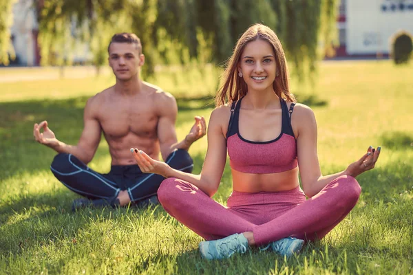 Healthy lifestyle concept. Happy young fitness couple meditating in lotus pose sitting on green grass. — Stock Photo, Image