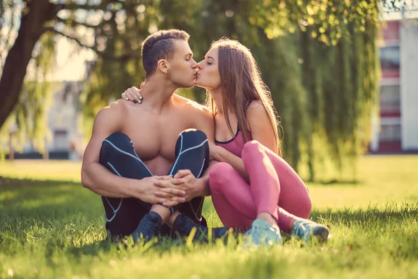 健康的生活方式概念。快乐的年轻健身情侣坐在绿色的草地上接吻. — 图库照片